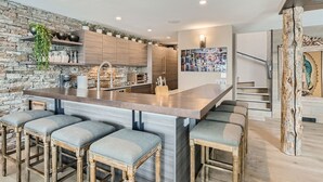 A modern kitchen with a central island, bar stools, a stone accent wall, and a staircase with a decorative railing. - 970-B Fairway Ct., Vail Golf Course Home - Hot Tub, Fire Pit,Gas Grill, Close to Lifts! (Vail)