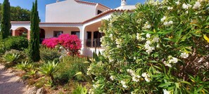 A white building with a tiled roof, surrounded by lush greenery and flowering plants.
