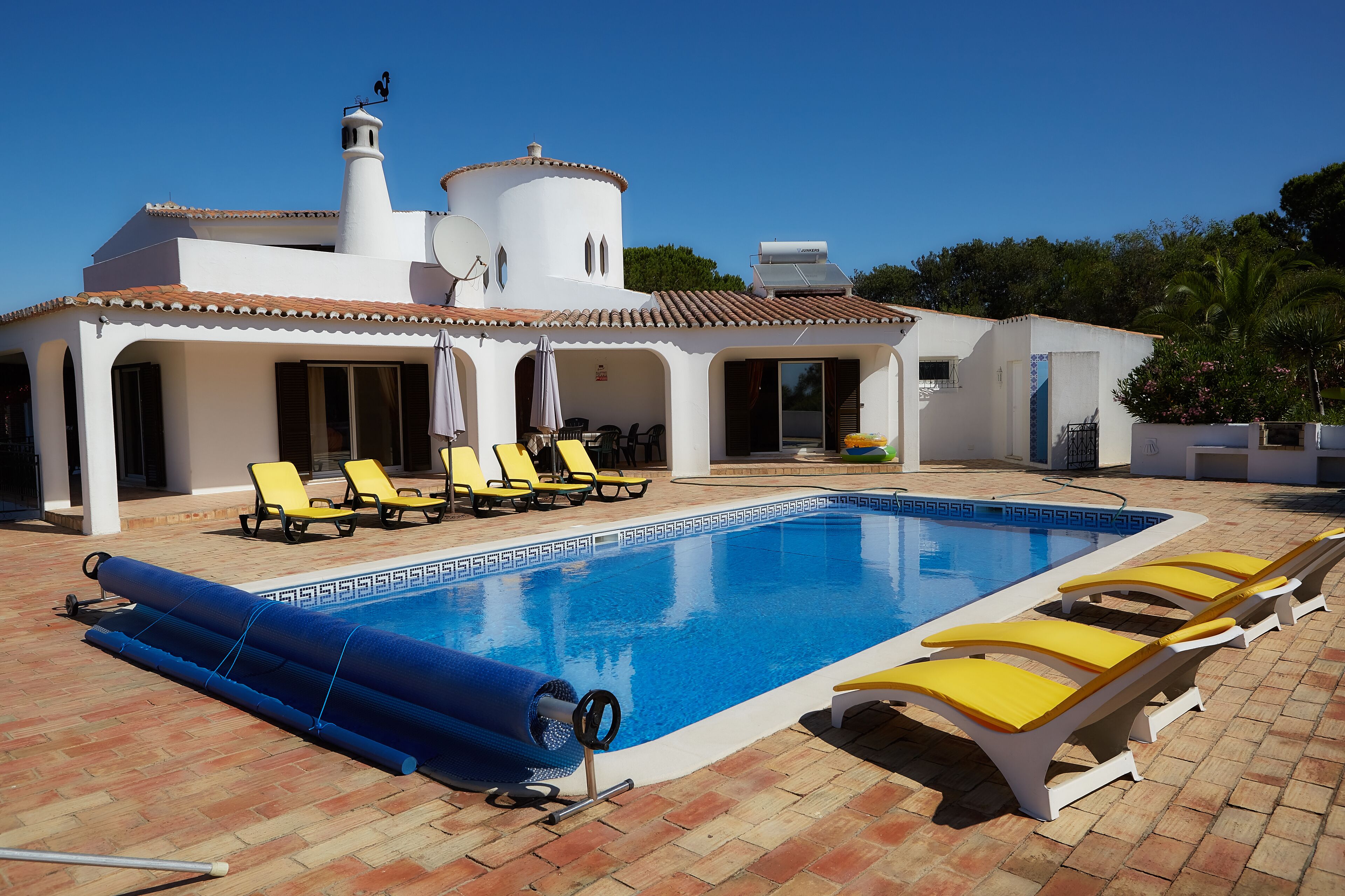 A swimming pool with sun loungers and a covered area, surrounded by a white building with a tiled roof.