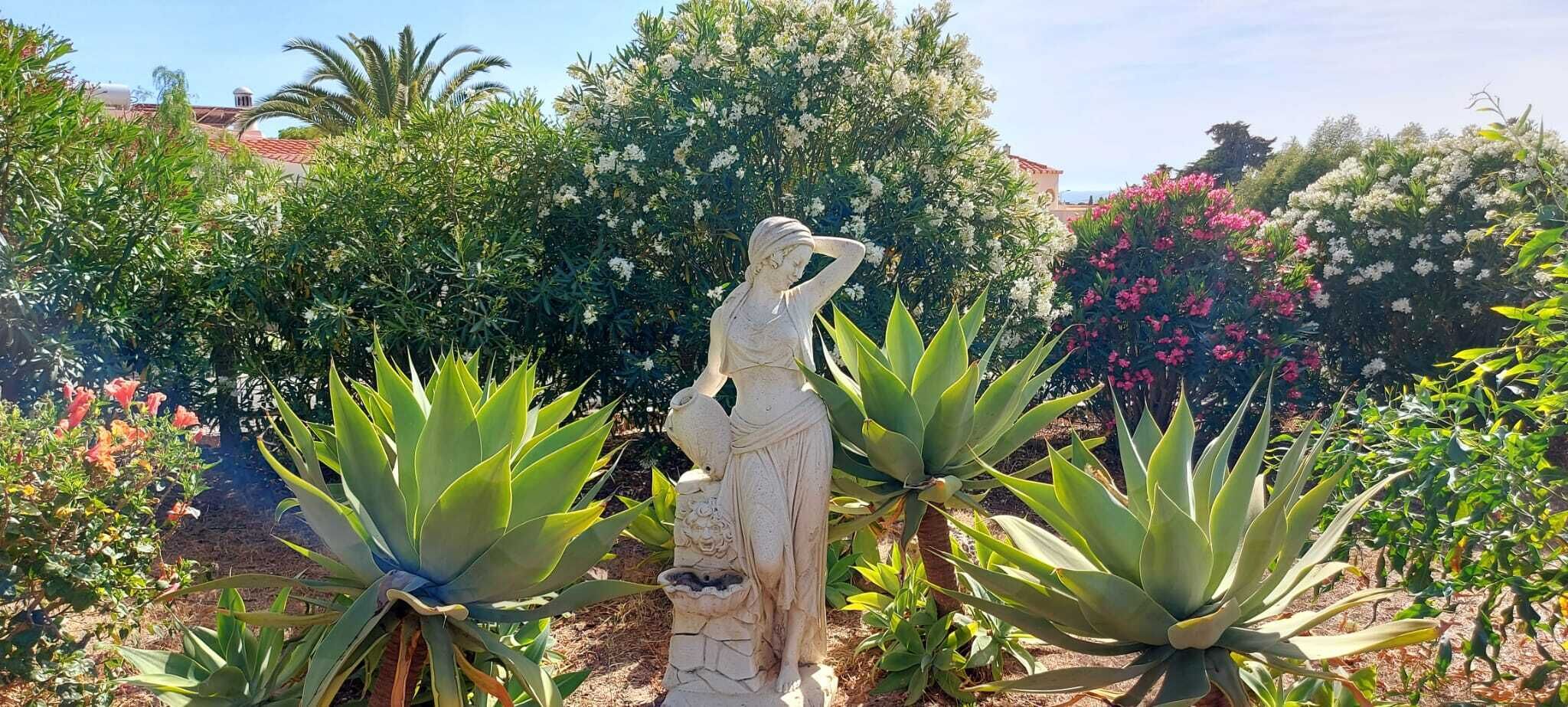 A garden with a statue, various plants, and a building in the background.