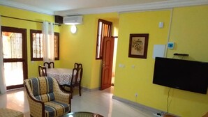 Una sala de estar con paredes amarillas, una mesa de centro de vidrio, un sofá, un televisor montado en la pared y un comedor con mesa y sillas.