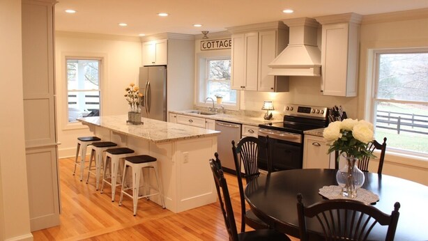 A modern kitchen with white cabinetry, a central island with bar stools, a dining table with chairs, and a view of the outside through a window.