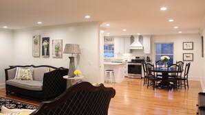 A modern living room with a sofa, a dining table with chairs, and a kitchen area with white cabinets and a stove.