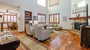 A spacious living room with hardwood floors, a fireplace, a bookshelf, and a dining area with wooden furniture. - Relax in Home w/ Htd. Pool+Hot Tub,  Putting Green, Whirlpool Bath+Steam Shower (Queen Creek)