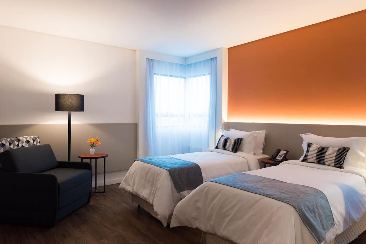 Ein Hotelzimmer mit zwei Betten, einem Schlafsofa, einem kleinen Tisch mit Blumenvase, einer Lampe und einem Telefon.