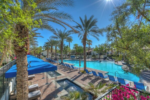 A pool area with lounge chairs and umbrellas, surrounded by palm trees and a clear blue sky.