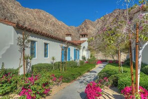 A white building with blue shutters, surrounded by lush greenery and pink flowers, with a mountainous backdrop.