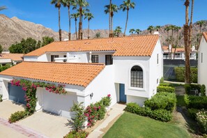 A white building with a red-tiled roof, surrounded by greenery and palm trees, with a mountainous backdrop. - Tennis Villa #3 - Courtside View, 2 bed, 2 bath, Permit 067145 (La Quinta)