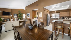 A modern kitchen with a central island, a dining area with a wooden table, and a living room with a fireplace. - Tennis Villa #3 - Courtside View, 2 bed, 2 bath, Permit 067145 (La Quinta)