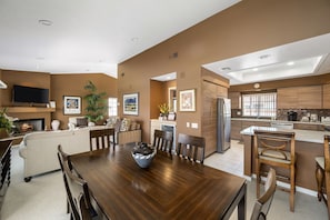 A modern kitchen with a central island, a dining area with a wooden table, and a living room with a fireplace. - Tennis Villa #3 - Courtside View, 2 bed, 2 bath, Permit 067145 (La Quinta)
