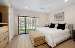A bedroom with a bed, a bench, a ceiling fan, a nightstand, a lamp, and a window with blinds. - Tennis Villa #3 - Courtside View, 2 bed, 2 bath, Permit 067145 (La Quinta)