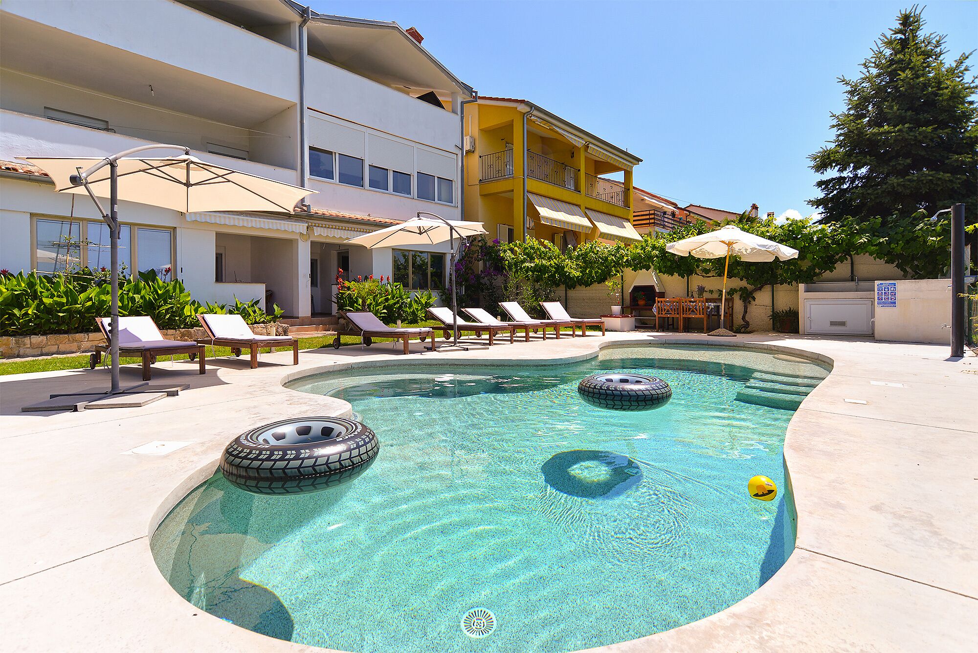 A swimming pool with lounge chairs and umbrellas, surrounded by a building with balconies and a clear sky.