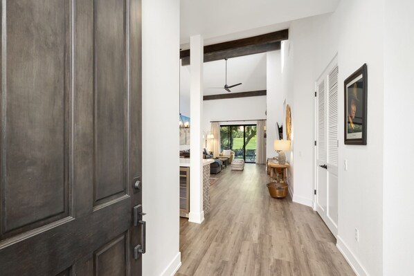 A hallway with a wooden floor, white walls, and a ceiling fan. There is a door on the left and a window on the right.