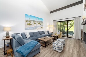 A modern living room with a grey sectional sofa, wooden coffee table, and striped ottoman. There are two side tables with lamps, a fireplace, and a large window with curtains.