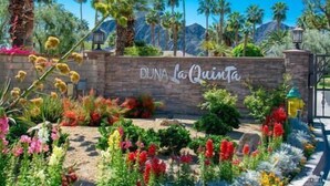 A sign with "DUNA La Quinta" written on a stone wall, surrounded by colorful flowers and palm trees. - Duna La Quinta #2 - Fairway Home - Pet Friendly (La Quinta)