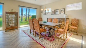 A dining room with a wooden table, matching chairs, a chandelier, and a large window with a view of a golf course. - Duna La Quinta #2 - Fairway Home - Pet Friendly (La Quinta)