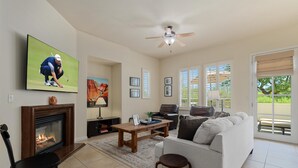 Una sala de estar con chimenea, televisor, ventilador de techo y un ventanal con persianas.