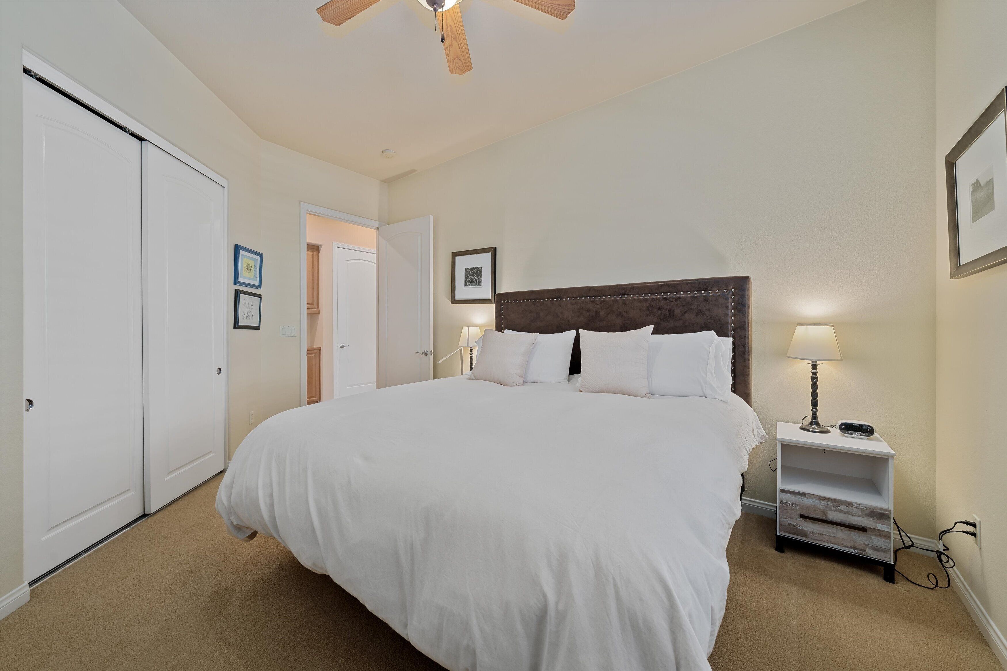A bedroom with a large bed, a nightstand with a lamp, a ceiling fan, and a door leading to another room.