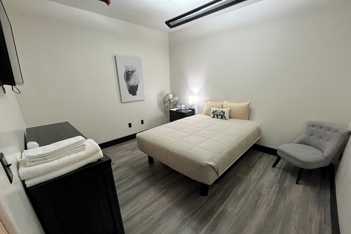 A bedroom with a bed, a chair, a bedside table with a lamp, and a wall-mounted picture.