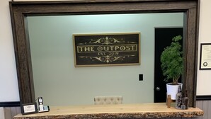Een receptiebalie met een 'WELCOME' bord erboven, een lijst met 'THE OUTPOST EST. 2019', een potplant en een ingelijst certificaat aan de muur.
