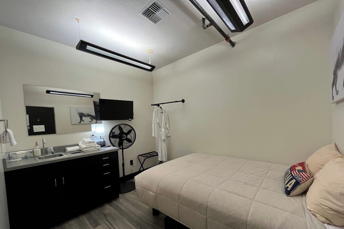 A modern bedroom with a bed, a bedside table, a mirror, a fan, and a TV.