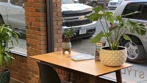 Een houten tafel met een zwarte stoel ervoor, voor een metselwerk muur met een raam. Op de tafel staan twee potplanten.