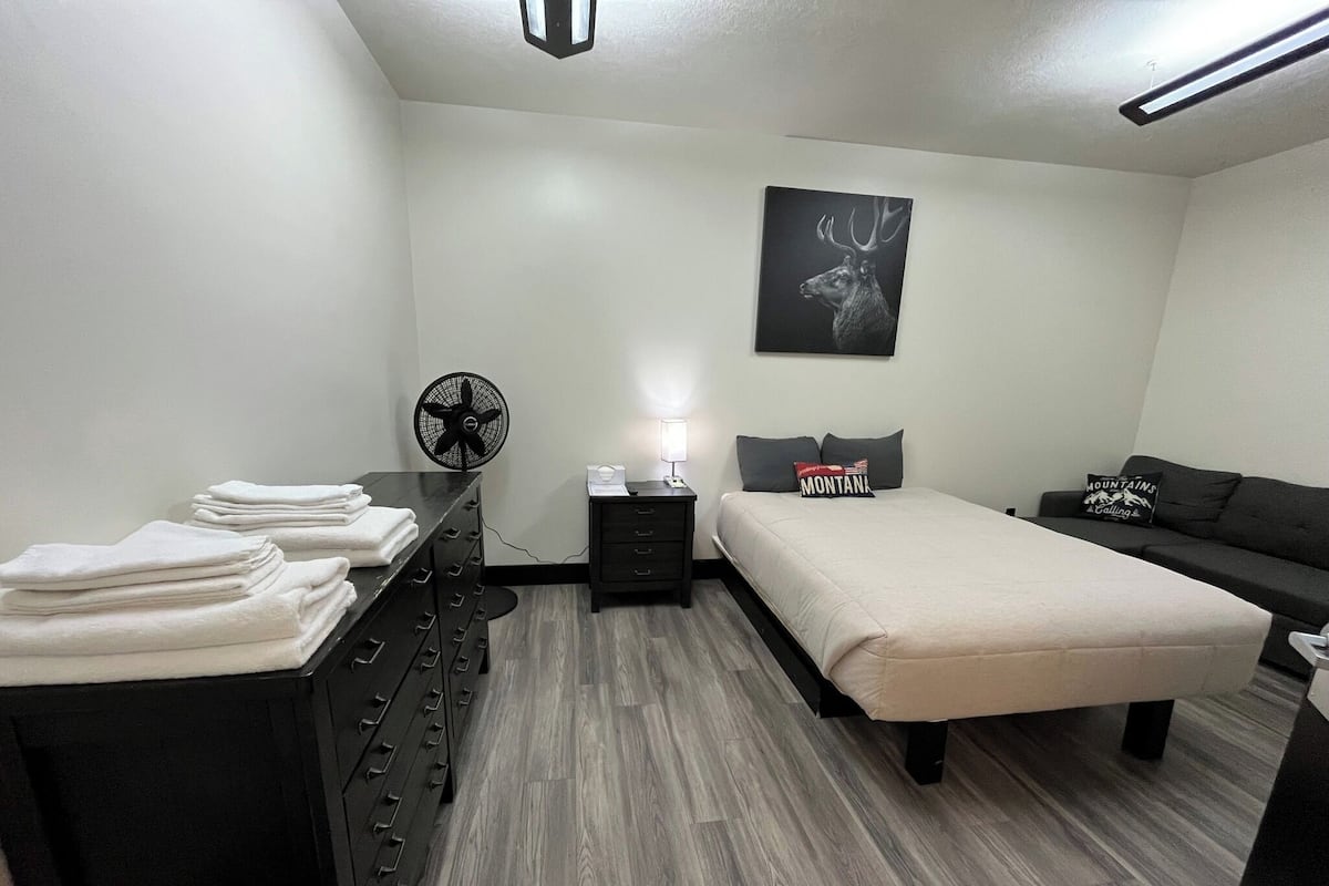 A modern bedroom with a bed, a dresser, a nightstand, a fan, and a wall-mounted artwork.