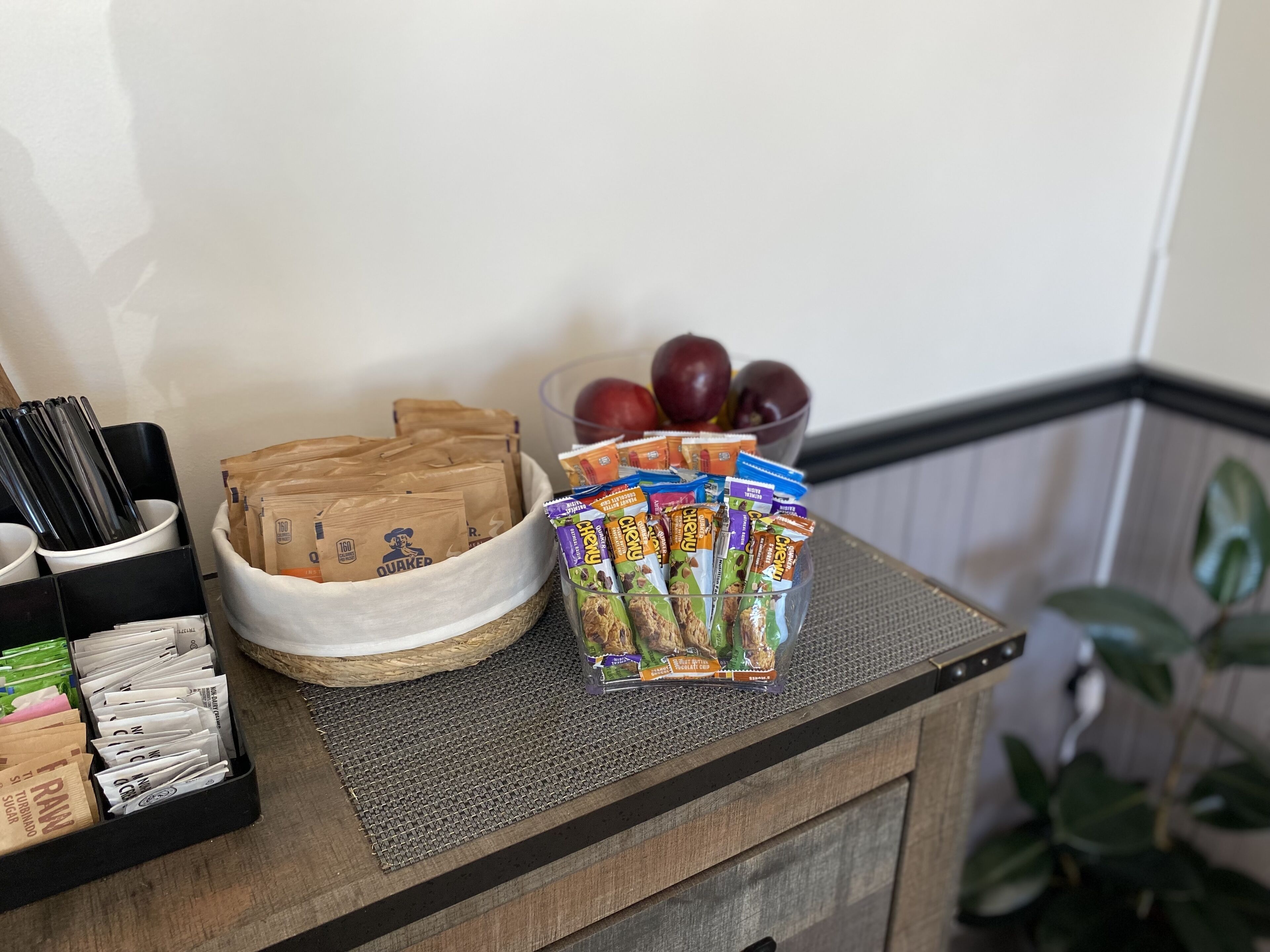 Una encimera de cocina con un cuenco de manzanas, una cesta de bolsas de papel marrón y un recipiente con paquetes de aperitivos.