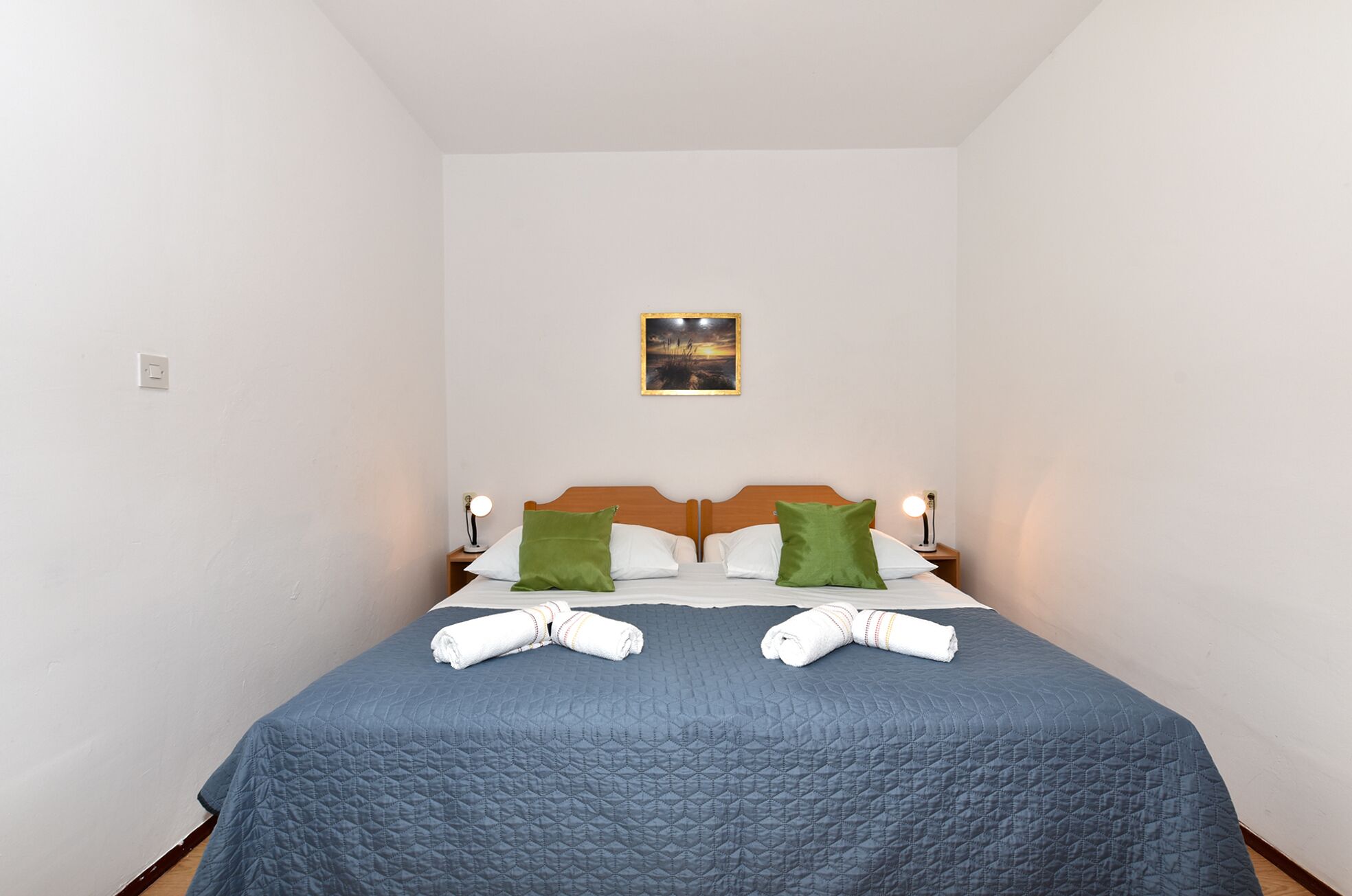 A neatly made bed with a blue quilt, two green pillows, and two white towels folded at the foot of the bed.