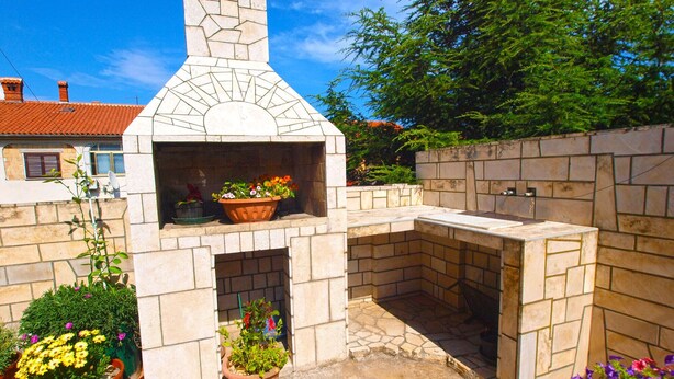 Un four en briques installé à l’extérieur, doté d’une cheminée, et entouré de plantes en pot et d’un mur de pierres.