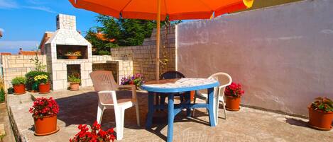 Une terrasse extérieure avec un barbecue en pierre, une table bleue, des chaises blanches et des plantes en pot.