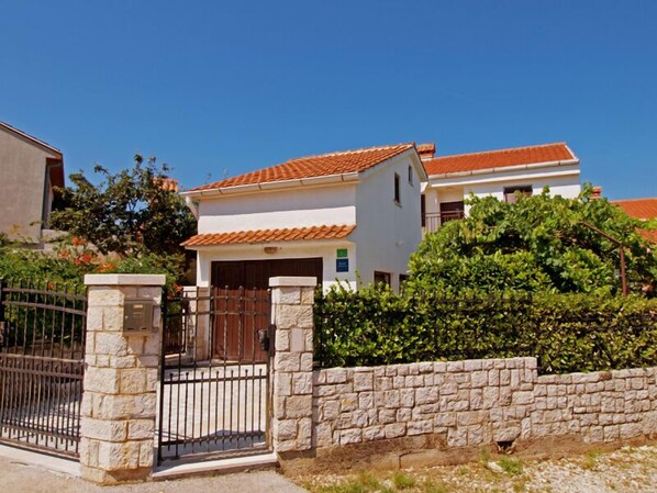 A two-story house with a tiled roof, a stone wall, and a metal gate.