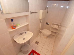A small bathroom with a white sink, toilet, and shower. There is a shelf with a soap dispenser and a toothbrush holder.
