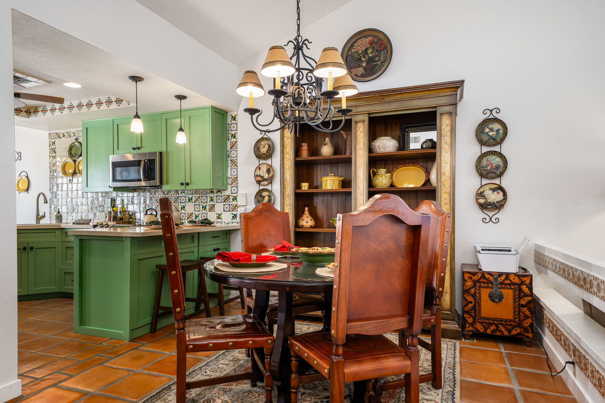A dining area with a round table, chairs, a chandelier, and a kitchen in the background.