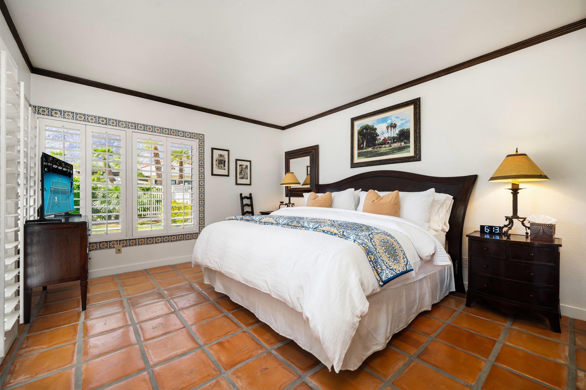 A hotel room with a bed, a nightstand, a lamp, a clock, and a window with a view of greenery.