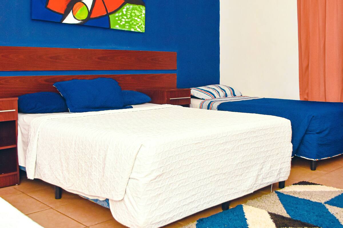 Un dormitorio con cama, dos mesitas de noche, una mesita auxiliar, un tapete azul y blanco estampado y un cuadro abstracto colorido en la pared.