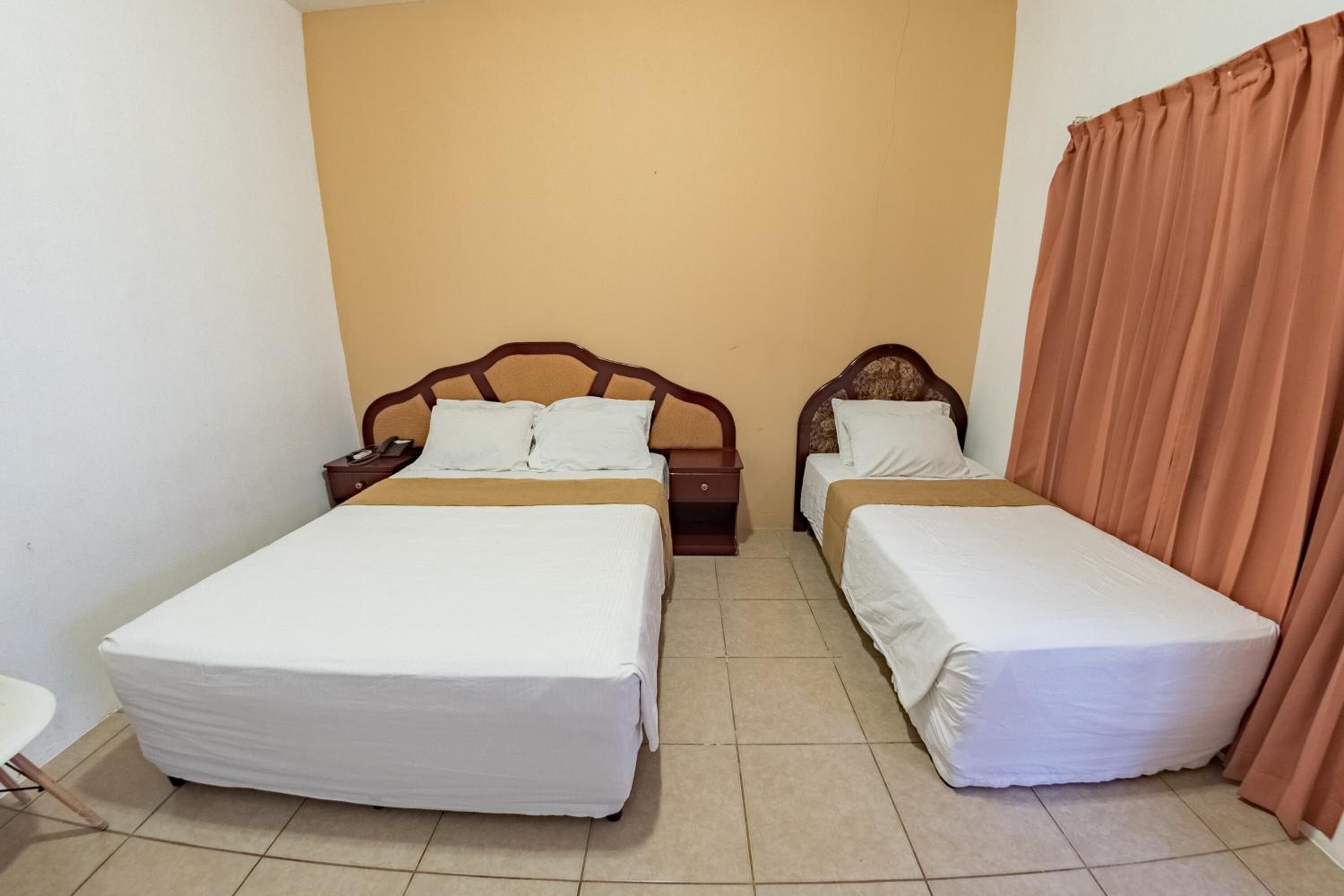 Habitación de hotel con dos camas individuales, cabecera de madera, mesita de noche y cortina marrón.