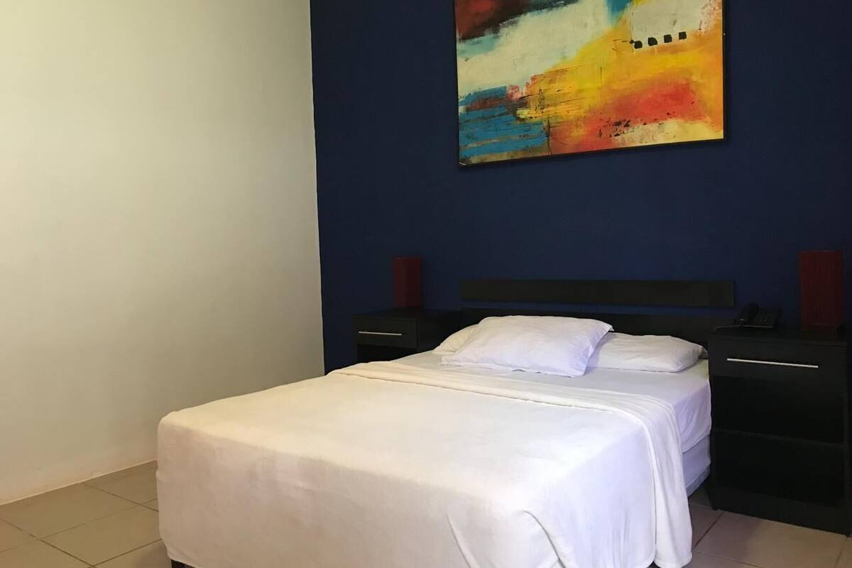 Una cama impecable con sábanas blancas en una habitación con una pared de acento azul oscuro y un cuadro abstracto colorido sobre ella.