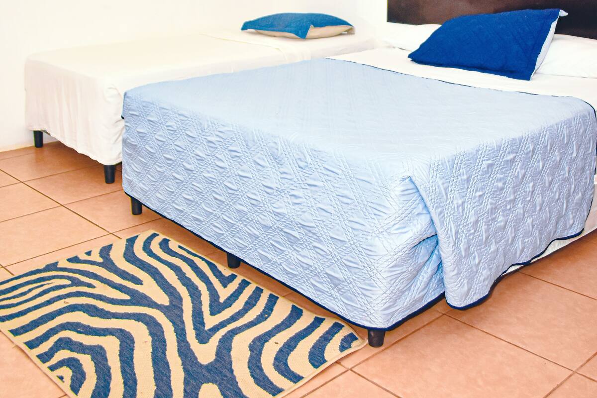 Un dormitorio con cama, una mesita de noche pequeña, un cojín azul, un tapete con estampado de cebra y un cuadro de colores en la pared.