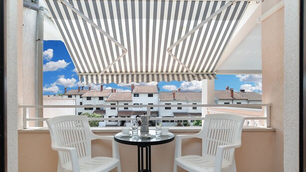 Un balcon avec deux chaises en plastique blanc et une petite table ronde, donnant sur une rangée d’immeubles aux toits de tuiles.