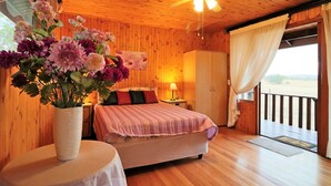 A wooden room with a bed, a nightstand, a vase of flowers, a ceiling fan, and a view through a window. - Tata Farm Game Lodge (Hammanskraal)
