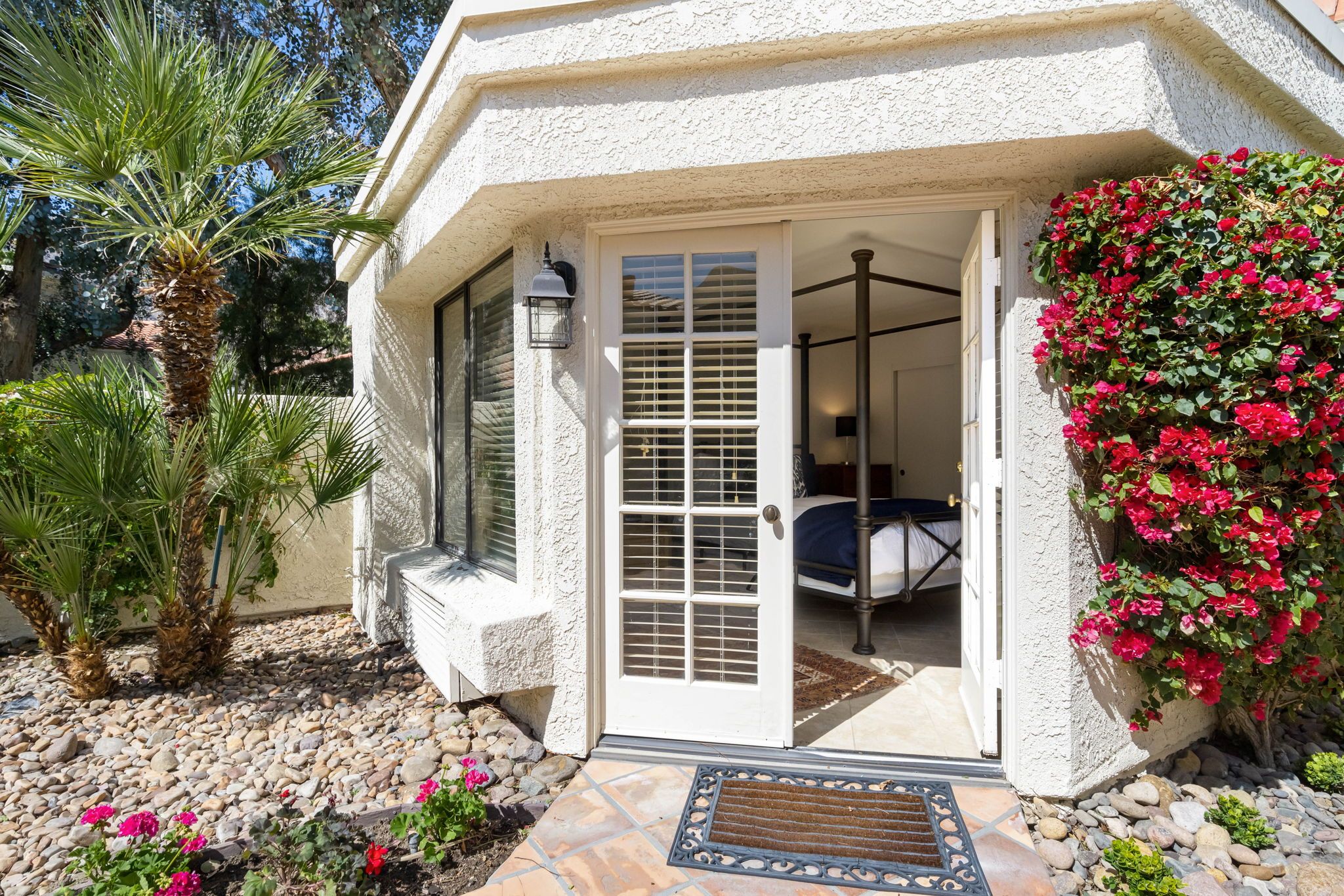 Uma casa com fachada branca, varanda coberta, porta com painel de vidro e pequeno jardim com pedras e plantas.