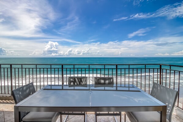 A table and chairs on a balcony overlooking a beach with a clear view of the ocean and sky.