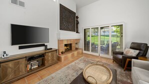 A living room with a flat-screen TV, fireplace, and a large window with blinds. - Santa Rosa Cove #14 Mtn. Views, 2 bed/2 bath Pet friendly Home, Permit 068343 (La Quinta)