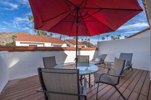 A patio with a glass table and chairs, a red umbrella, and a view of buildings and palm trees. - Tennis Villa #35 - Courtside Views, 2 bedroom, 2 bathroom, permit 063433 (La Quinta)