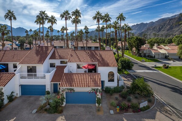 A residential area with houses featuring red-tiled roofs and white walls, surrounded by palm trees and mountains in the background.