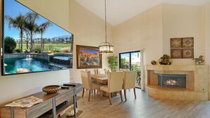 Amplia sala de estar con un televisor grande mostrando una vista panorámica, una chimenea y un comedor con una mesa y sillas de madera.