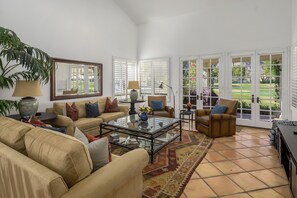 A spacious living room with a beige sofa, a glass coffee table, and a brown leather armchair. There is a large window with white shutters, a potted plant, and a vase with flowers on the coffee table. - Santa Rose Cove #41, 3 BD/ 3.5 Bath, Pet Friendly Home (La Quinta)