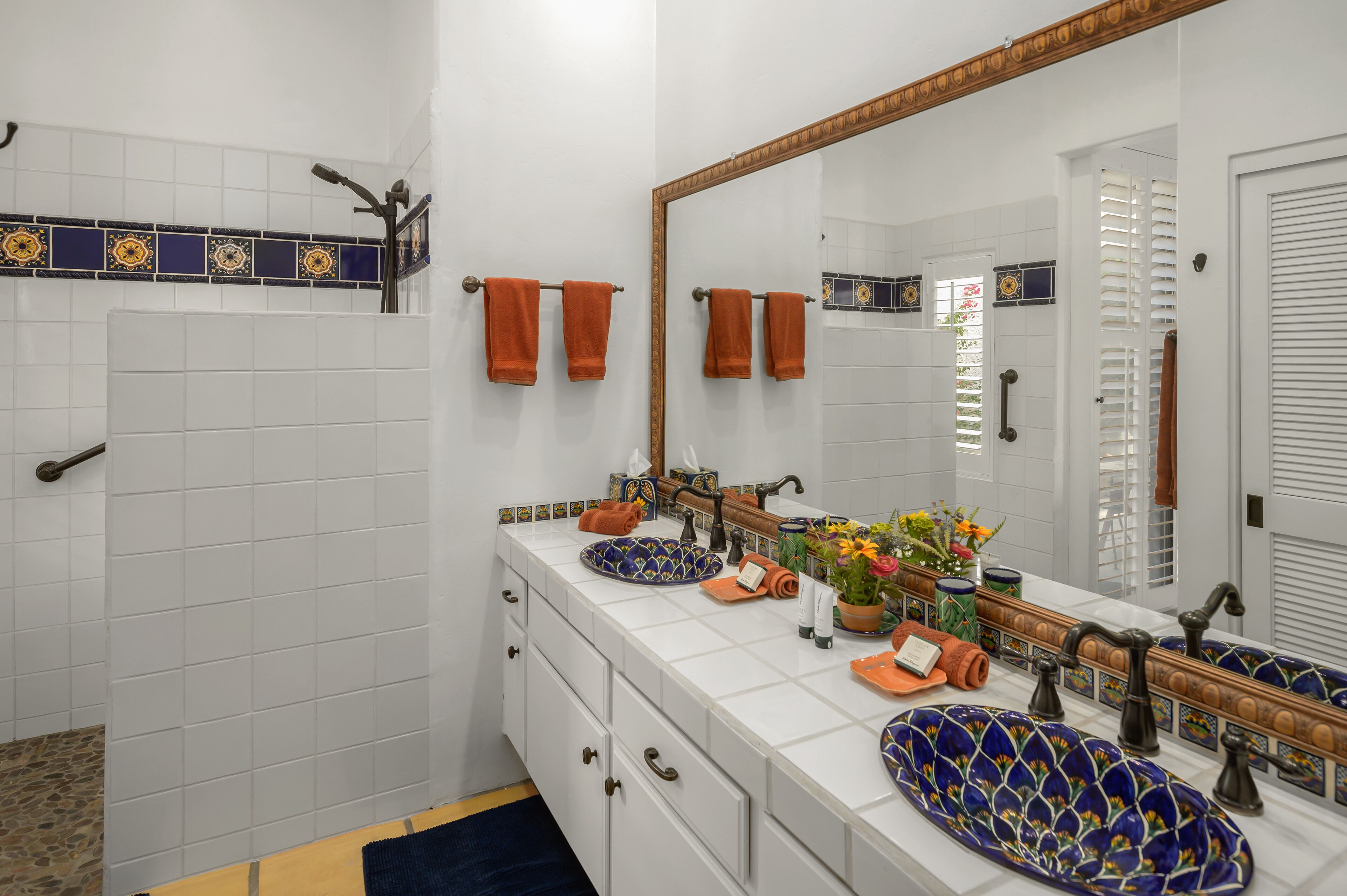 Une salle de bain aux murs recouverts de carreaux blancs, avec un grand miroir, deux vasques ornées de jolis récipients et des serviettes rangées sur un porte-serviettes.