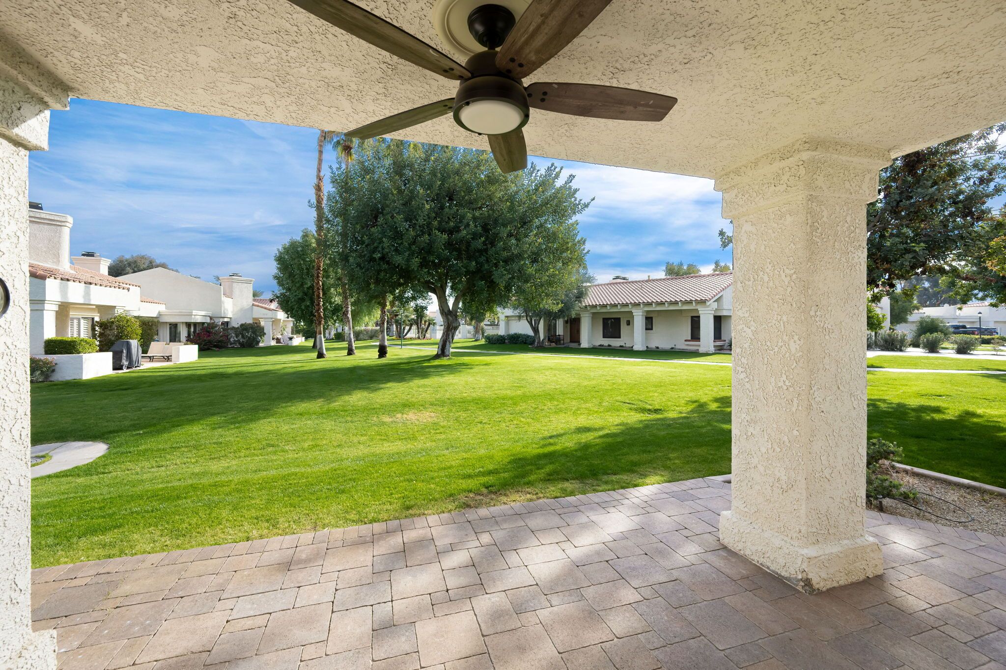 A covered patio area with a ceiling fan, a well-maintained lawn, and a view of residential buildings in the background.